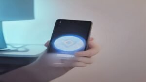 Mão segurando um celular Android com gráficos coloridos de figurinhas na tela em uma mesa de trabalho moderna.